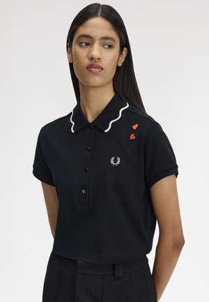 Mujer con cabello largo y oscuro, vestida con una camiseta tipo polo negra con ribete blanco en el cuello y dos parches en forma de corazón rojo en el hombro.
