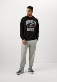 Felpa nera con grafica "BROOKLYN NETS 2012" in bianco e dettagli floreali, abbinata a pantaloni della tuta grigi e scarpe da ginnastica scure.