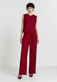 Top rosso senza maniche con un collo alto e due bottoni neri, abbinato a pantaloni a gamba larga, entrambi in un tessuto morbido.