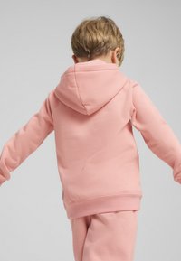 Rozā kapuces džemperis, izgatavots no mīkstas auduma, ar brīvu piegriezumu un ribotām aprocēm. Aizmugures skats atklāj gludu tekstūru bez uzdrukām.