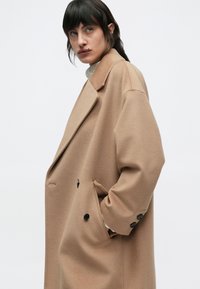 Bézs színű, túlméretezett kabát, puha, vastag anyagból készült. Jellemzői a csapott gallér, a dupla soros gombolás és a fekete gombdíszítés.