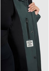 Duga zelena pufasta jakna otvorena, s crnim unutrašnjim dijelom. Sadrži zippered džep i oznaku koja kaže: "Topli zagrljaj od tkanine."