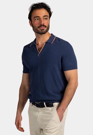 Man met baard die een donkerblauw poloshirt met korte mouwen en oranje bies draagt en crème broek heeft, staat met één hand in zijn zak, kijkt naar de zijkant.