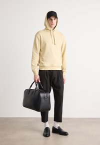 Felpa beige con cappuccio, pantaloni neri, borsa in pelle nera, calzini bianchi e mocassini neri. Texture morbide, design minimalista.