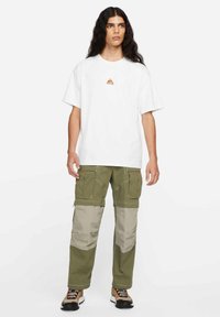 White t-shirt with a small orange logo, paired with green cargo pants featuring beige panels, multiple pockets, and a relaxed fit.
