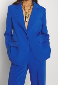 Blauwe op maat gemaakte blazer met een enkele knoop, geknipte revers en twee voorzakken, gecombineerd met gouden kettingjuwelen en een diepe halslijn.