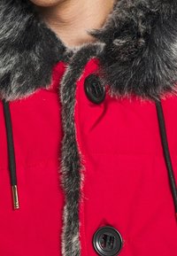 Rød pufferjakke med en krave af falsk pels i mørkegrå, med sorte knapper og snore. Stoffet har en glat tekstur.