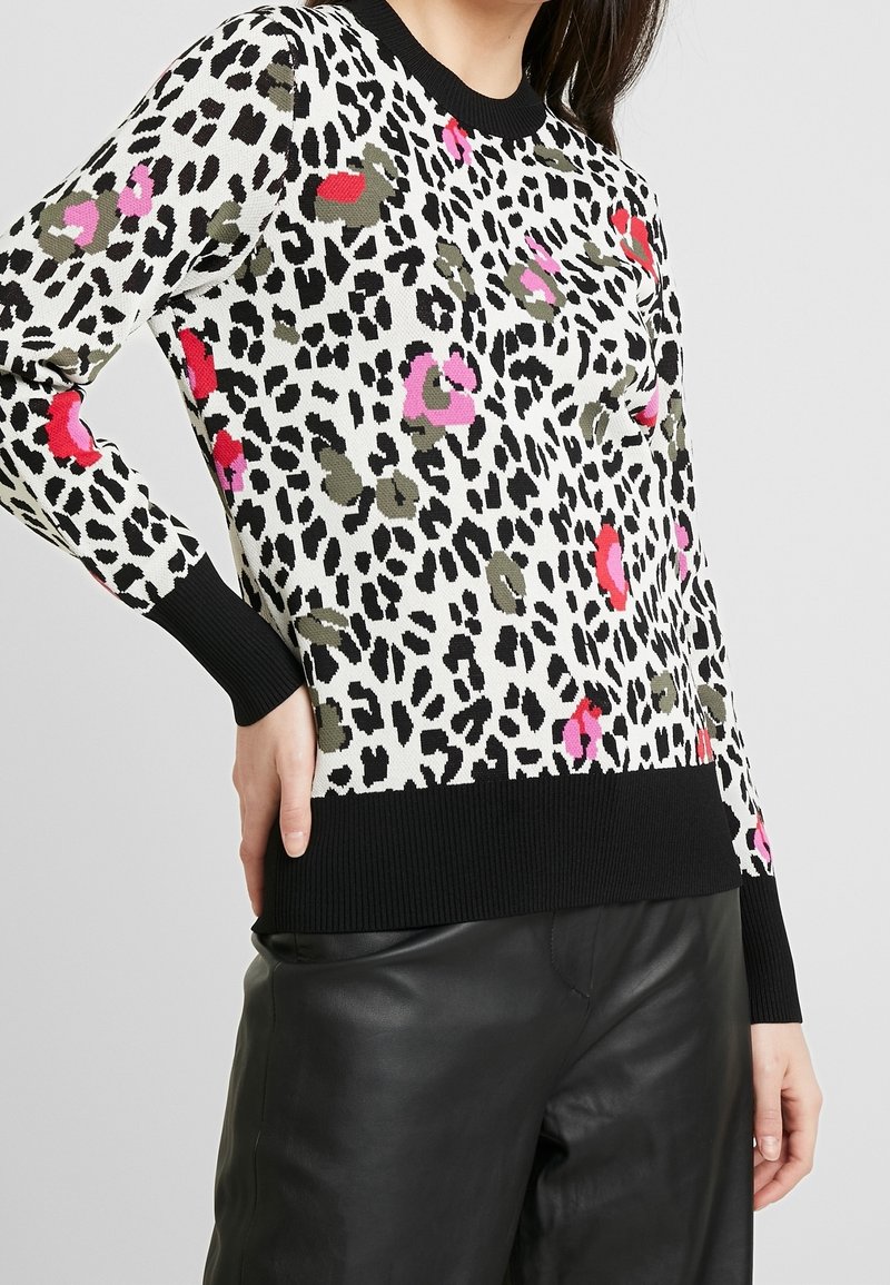 Femme portant un pull blanc avec imprimé léopard noir, rose, rouge et olive, bords-côtes noirs aux poignets et à l'ourlet, associé à un pantalon en cuir noir.