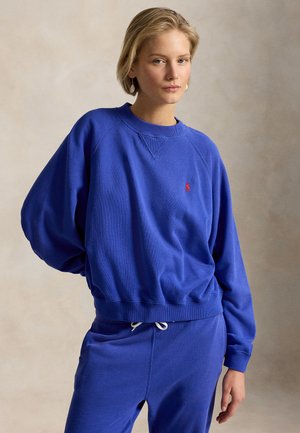 Noor naine lühikeste blondide juustega, kellel on seljas kuningsinine dressipluus ja sobivad dressipüksid, seismas neutraalse tausta ees.