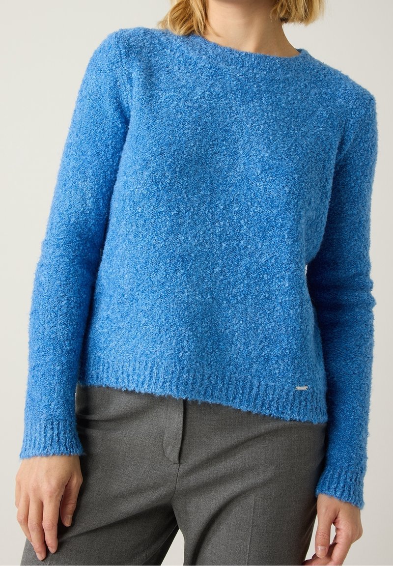Femme portant un pull texturé bleu vif à manches longues associé à un pantalon gris sur mesure, debout devant un fond uni.
