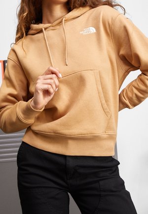 Kobieta w beżowej bluzie z kapturem The North Face z przednią kieszenią i czarnych spodniach, pozująca z jedną ręką przy kieszeni, a drugą na talii.