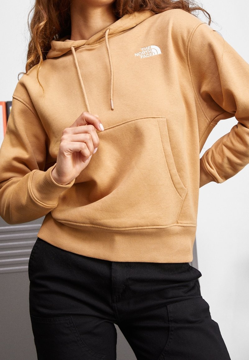 Femme portant un sweat à capuche beige The North Face avec une poche avant et un pantalon noir, posant avec une main près de la poche et l'autre à la taille.
