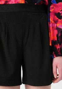 Sorte shorts laget av mykt materiale, med folder, flat linning og en nederkant som slutter over knærne. Synlige sømdetaljer.