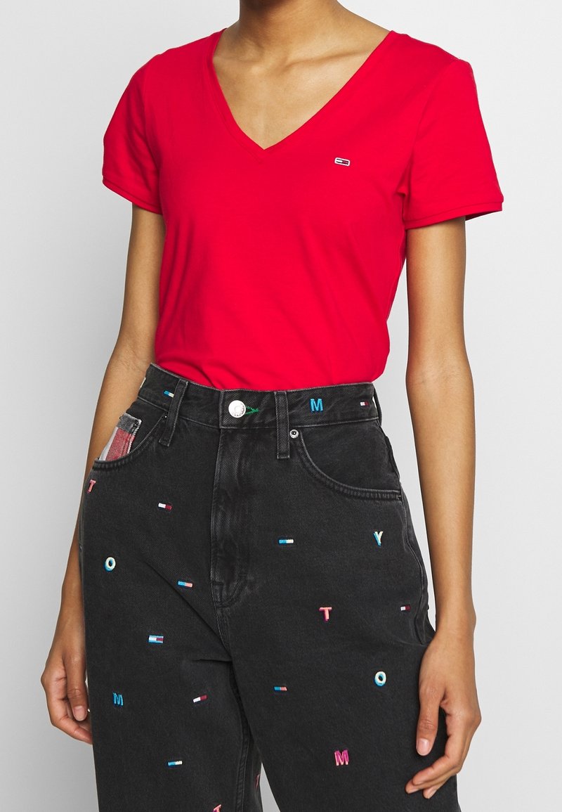 Personne portant un T-shirt rouge à col en V et un jean noir taille haute avec des lettres brodées multicolores et de petits logos de drapeaux.