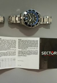 Orologio in acciaio inossidabile argentato con quadrante nero testurizzato, accento blu sulla ghiera e bracciale metallico. Include libretto di garanzia.