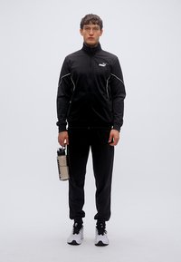 Μαύρη αθλητική φόρμα με ζακέτα με φερμουάρ και λευκές λεπτομέρειες, ταιριαστό παντελόνι και λευκά αθλητικά παπούτσια. Κρατάει μια διάφανη πλαστική φιάλη νερού.