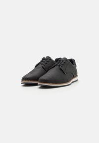Chaussures noires texturées avec une surface perforée, design à lacets et semelle contrastante blanche et tan. Forme mince avec un bout arrondi.