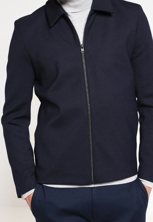 Homme portant une veste zippée bleu marine foncé sur un pull à col roulé gris clair, les mains dans les poches du pantalon bleu marine.