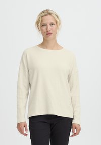 Maglione a maniche lunghe color beige chiaro a costine con scollatura ampia e vestibilità comoda. Indossato con pantaloni neri, mette in risalto una consistenza morbida e un design rilassato.
