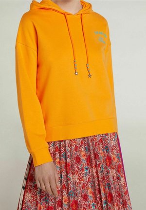 Personne portant un sweat à capuche orange avec des perles en forme d'étoile sur le cordon de serrage et une jupe plissée rouge à motifs floraux sur un fond uni.