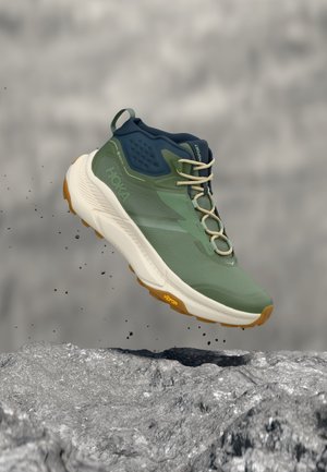 Vihreä ja laivastonsininen vaelluskenkä, jossa valkoinen pohja ja Vibram-kuvio, näkyy ilmassa kivikkoisen maan yläpuolella, pölyn ja pienten kivien pudotessa.