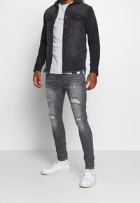 Chaqueta de mezclilla negra con bolsillos, jeans grises rasgados y zapatillas blancas. El conjunto presenta un estilo en capas con una camiseta gris debajo.