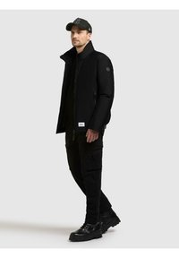 Black jacket featuring a high collar, zip closure, and side pockets. Styled with black cargo pants and sturdy black boots.