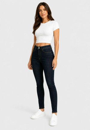 Kvinde med langt brunt hår, iført en hvid crop top, mørke skinny jeans og hvide sneakers, står mod en ensartet lys baggrund.