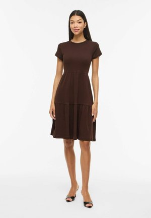 Kobieta stoi, ubrana w ciemnobrązową sukienkę z krótkim rękawem sięgającą do kolan z warstwową spódnicą oraz czarne sandały na obcasie na białym tle.