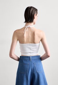 Weißes Halter-Crop-Top mit einer Schleife am Hals, kombiniert mit hochgeschnittenen blauen Denim-Hosen, die zwei Gesäßtaschen haben.