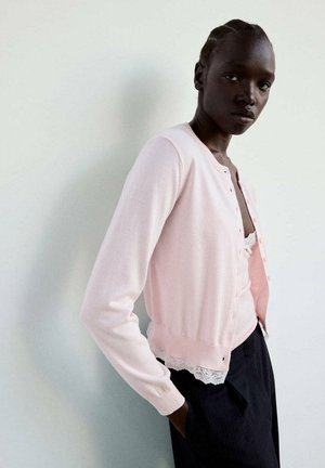 Femme à la peau foncée et aux cheveux tressés portant un cardigan rose clair par-dessus un top orné de dentelle et un pantalon foncé, debout contre un mur blanc uni.