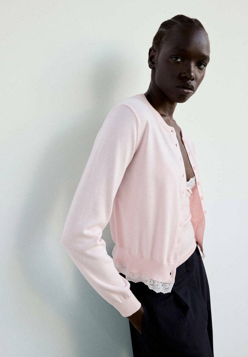 Femme à la peau foncée et aux cheveux tressés portant un cardigan rose clair par-dessus un top orné de dentelle et un pantalon foncé, debout contre un mur blanc uni.