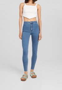 Höga blåjeans, elastisk denimtyg, slim fit. Kombinerade med en vit ribbad crop top och gröna sandaler med spännen.