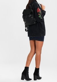 Zaino in pelle nera con pattina ripiegabile, dettagli in metallo dorato, spallacci regolabili e una texture liscia. Design di rosa ricamata sulla giacca.