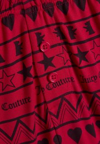 Piros anyag fekete csillag-, szív- és medvemintákkal. Két rózsaszín gomb és rugalmas derékrész. A szöveg "Juicy Couture" feliratot tartalmaz.