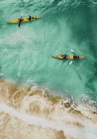 Two people each paddling yellow kayaks in turquoise water near a sandy shore with white waves.