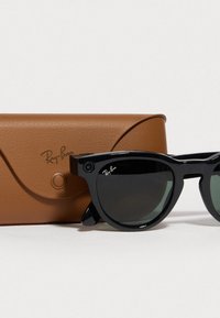 Occhiali da sole neri con lenti scure, caratterizzati da un design audace e arrotondato e due piccole telecamere sulla parte anteriore, accanto a un astuccio in pelle marrone.