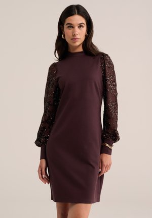 Femme portant une robe violet foncé longueur genou avec des manches longues transparentes et pailletées, des boucles d'oreilles créoles dorées et un bracelet, debout devant un fond uni.