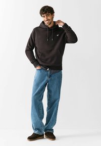 Ciemnobrązowa bluza z kapturem, z kieszenią kangurką, noszona na paskowanej koszuli; zestawiona z luźnymi niebieskimi dżinsami i ciemnobrązowymi butami wsuwanymi.