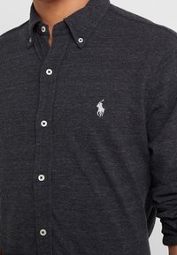 Mörkgrå skjorta med knappar i vitt och en liten vit broderad polo-spelare-logotyp på vänstra bröstet.