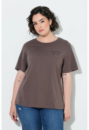 Kvinde med kort krøllet hår iført en brun T-shirt med teksten "Irresistibly Yours" og blå jeans, stående mod en hvid væg.