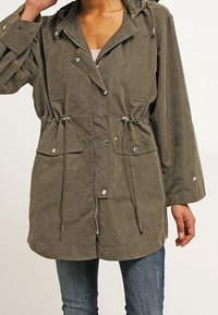 Olivově zelená bunda s kapucí, stahovacími šňůrkami, patentkami, dvěma předními kapsami a volným střihem, vyrobená z měkké, texturované tkaniny.