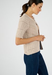 Cardigan en maille beige à manches courtes, présentant des motifs ajourés, un col rond et une fermeture à boutons. Associé à un jean bleu foncé.
