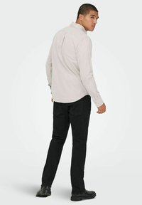 Beige langærmet skjorte med knap-manchetter, rundet kant og rygdetalje, parret med sorte slim-fit bukser og sorte sko.