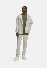 Hellgraue Kapuzenjacke über einem grünen T-Shirt, kombiniert mit hellgrauen Hosen und weißen Sneakers. Verfügt über elastische Bündchen und einen Reißverschluss vorn.