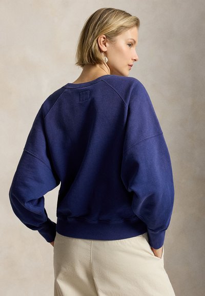 Γυναίκα με κοντά ξανθά μαλλιά, που φοράει μια φαρδιά μπλε σκούρα φούτερ και μπεζ παντελόνι, στέκεται με τα χέρια στις τσέπες και κοιτάζει στο πλάι.