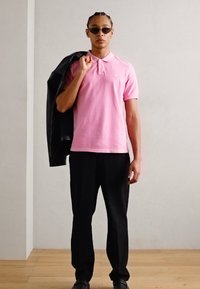 Rosa polo-shirt med korte ærmer, sorte bukser og en sort læderjakke hængende over den ene skulder. Runde solbriller fuldender looket.