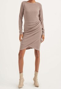 Abito aderente a maniche lunghe in taupe chiaro realizzato in tessuto a maglia testurizzata. Dettaglio drappeggiato in vita, abbinato a stivali alla caviglia beige.