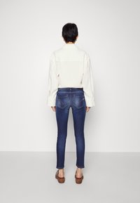 Camisa blanca de manga larga con cuello, combinada con unos vaqueros ajustados de color azul oscuro que tienen bolsillos traseros y detalles de costura. Sandalias de plataforma marrones.