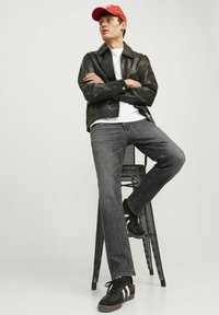 Veste en cuir noire, t-shirt blanc, jean gris et baskets noires avec des rayures blanches. Casquette rouge portée à l'envers. Assis sur un tabouret en métal.